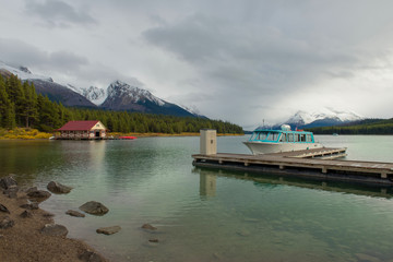 Fototapeta na wymiar Lago maligne chalet que se ubica en Jasper Alberta Canada, un lago hermoso en medio de las montañas, una cabaña roja, un muelle y un pequeño barco azul en un cielo nublado y un lago azul turquesa