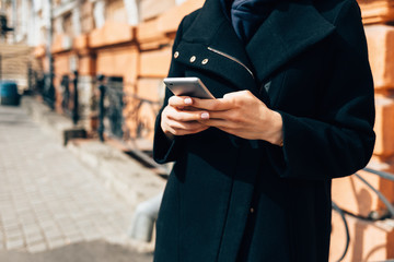 Close-up young woman wearing elegant black coat holding mobile phone standing on street in old European city in daytime.