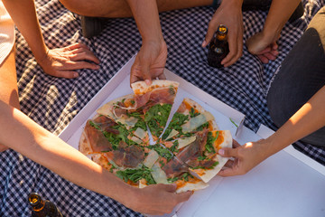 Group of friends eating pizza outdoors. Man and women sitting on plaid, taking pizza slices and drinking beer. Picnic concept