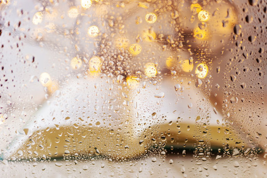 An open book lies against a light wall with a garland behind a glass covered with raindrops. The book is blurred, the focus is glass with drops. Reading concept in the rain, cozy still life.