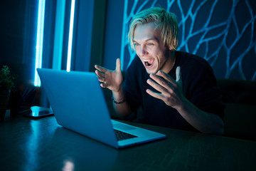 A young guy player sits at a table with a laptop and yells from winning an online game against a background of blue neon