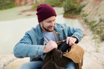 Bearded hipster in jeans pulls out of a backpack portable speaker to listen to music in nature
