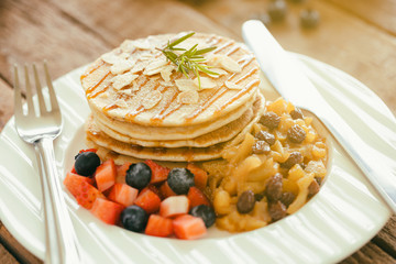 Homemade pancake or hot cake stacked on white plate with fresh fruit and apple raisin sauce on wood table. Pancakes for breakfast or dessert with caramel sauce and sugar icing so sweet and delicious.