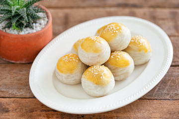 Chinese flaky pastry or moon cake filled with sweet mung bean paste and salted egg yolk on white plate decorated with white sesame in top view with copy space on wood table. Homemade bakery concept.