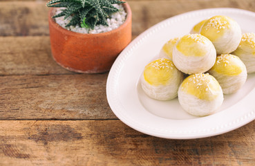 Chinese flaky pastry or moon cake filled with sweet mung bean paste and salted egg yolk on white plate decorated with white sesame in top view with copy space on wood table. Homemade bakery concept.