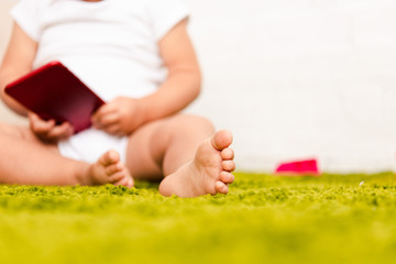 Partial view of little barefoot child sitting on green floor and holding digital device