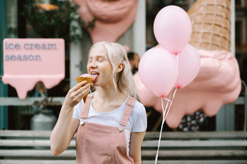 Cute girl with blond hair licks ice cream with her eyes closed and enjoys smiling, holding balloons...