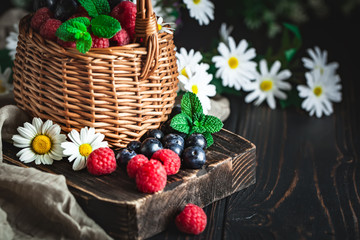 Fototapeta na wymiar Raspberries and blueberries in a basket with chamomile and leaves on a dark background. Summer and healthy food concept. Background with copy space. Selective focus.