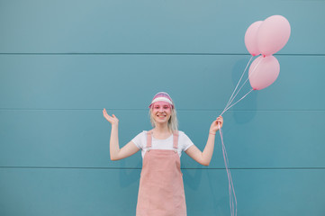 Cheerful girl with balloons in her hand stands on a blue background in pink clothes, looks into the camera, smiles and spreads her hands to the sides. Isolated on wall background.