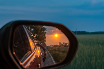 Look in the outside mirror of a moving car. In the mirror you can see the setting sun, parts of the vehicle and a bicycle at the rear.