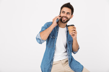 Image of handsome joyful man drinking coffee takeaway and talking on cellphone while sitting
