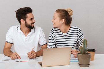 Loving couple friends man and woman sitting at the table indoors isolated over grey background using laptop computer reading book studying.