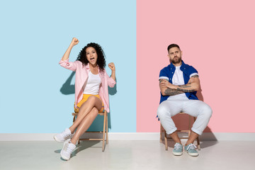 Obraz premium Young emotional caucasian couple in bright casual clothes posing on pink and blue background. Concept of human emotions, facial expession, relations, ad. Woman celebrating, man is bored and sad.