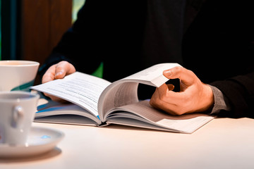 Close up of man reading book and drinking tea or coffee on rainy day in cozy modern cafe.