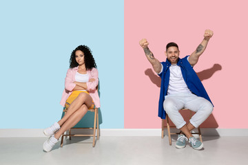 Young emotional caucasian couple in bright casual clothes posing on pink and blue background. Concept of human emotions, facial expession, relations, ad. Woman bored and sad, man celebrating, happy.