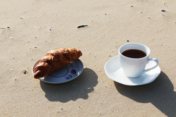 A cup of coffee with a croissant stands on the seashore