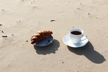 A cup of coffee with a croissant stands on the seashore