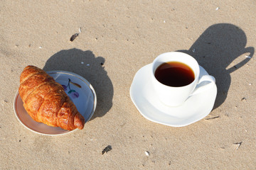 A cup of coffee with a croissant stands on the seashore