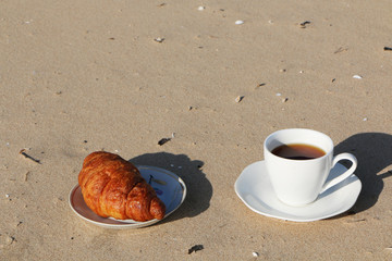 A cup of coffee with a croissant stands on the seashore