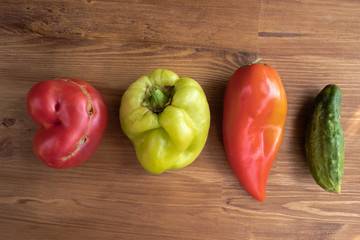 Ugly vegetables: red tomato, green sweet pepper, red sweet pepper, cucumber on a brown board, top view
