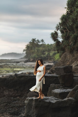 Young pregnant woman in white dress enjoying evening in Bali beach. Sensitivity to nature and harmony. Maternity concept.