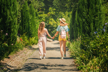 Naklejka na ściany i meble Happy girl friends walking by a path in local park among trees at summer day