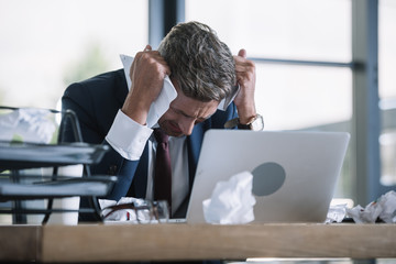 selective focus of upset man in suit holding crumpled paper near laptop