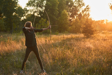 A woman shoots a bow in nature at sunset.