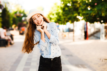 Emotional girl teenager in a hat walks through the summer city. Debonair caucasian girl enjoying photoshoot on city background.