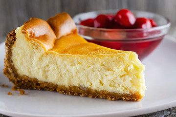 A slice of New York style cheesecake with a bowl of cherry sauce blurred in the background.  White plate.  Neutral background.