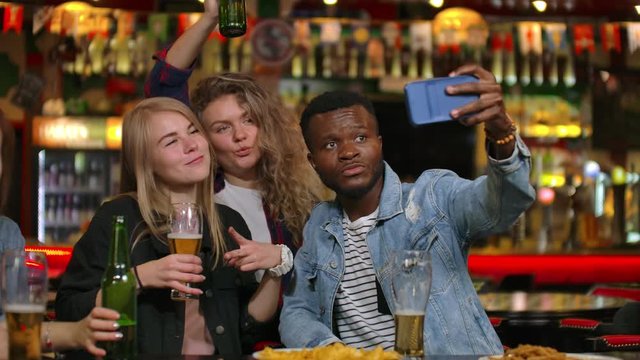 A group of friends multiethnic resting in the bar. Friends take a photo on the phone at the bar, make a shared photo on the phone. Party with friends at the bar with beer