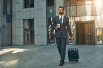 Busy morning. Full length of young and handsome bearded businessman in suit pulling his luggage and holding cup of coffee while walking outdoors