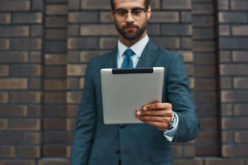 Modern businessman. Portrait of handsome bearded businessman in eyeglasses working with touchpad while standing against brick wall