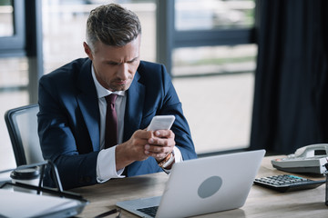 handsome businessman using smartphone near laptop and document tray