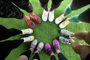 Closeup of athletes wearing sports shoes and standing in circle on green grass, top view. Space for text - Powered by Adobe