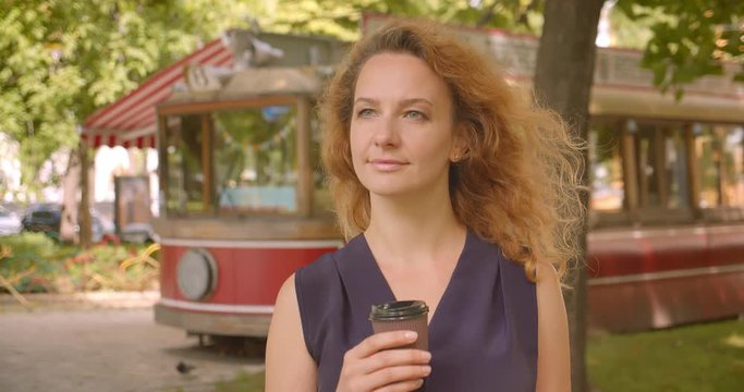 Closeup portrait of adult pretty gingerhead female looking at camera smiling cheerfully holding coffee in park outdoors
