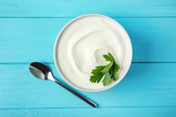 Bowl of fresh sour cream with parsley and spoon on light blue wooden table, flat lay