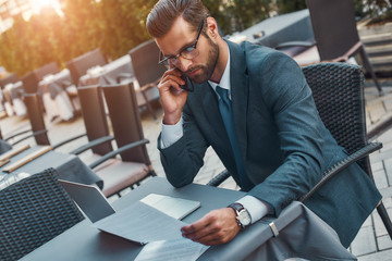 Working from everywhere. Portrait of handsome bearded businessman in eyeglasses talking by phone with client and looking at documents while sitting in restaurant outdoors