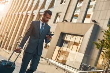 Always available. Young and handsome bearded man in suit pulling suitcase and looking at his smartphone while walking outdoors