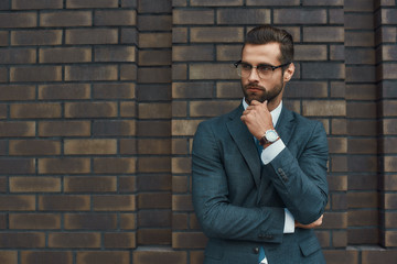Lost in thoughts. Portrait of handsome bearded businessman in eyeglasses and formal wear thinking about something while standing against brick wall