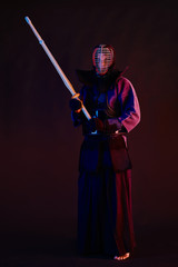 Fototapeta premium Bliska strzał, wojownik Kendo ubrany w zbroję, tradycyjne kimono, hełm uprawiający sztukę walki z bambusowym mieczem shinai, czarne tło.