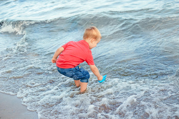 Happy Caucasian red-haired toddler child boy putting blue paper boat in water on lake sea ocean shore at evening sunset or morning. Kid playing on beach. Happy lifestyle childhood concept.