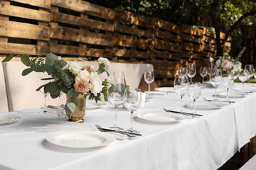 Wedding table setting decorated with fresh flowers in a brass vase. Wedding floristry. Banquet table for guests outdoors with a view of green nature. Bouquet with roses, eustoma and eucalyptus leaves
