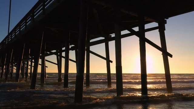 Aerial view under pier at Newport Beach as waves roll in as the sun begins to set.