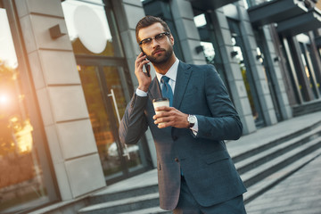 Important talk. Young and handsome bearded businessman in formal wear talking by phone and holding cup of coffee while standing outdoors