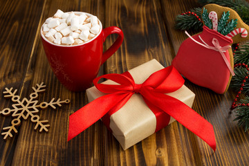 Christmas gift with red  ribbon, hot chocolate with marshmallows in the red cup and gingerbreads  on the brown wooden background