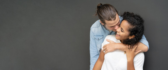 Panoramic banner image of a sweet couple hugging and kissing his wife on the cheek on black background in studio. wide crop - Powered by Adobe