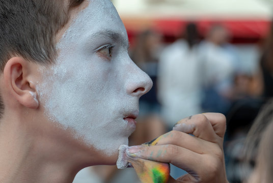 Theatrical performance on the streets. A female hand applies white makeup to the face of a teenager boyfriend. The boy in the image of a clown or mime.