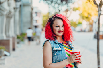 Stylish happy young woman in a bright striped sundress and a denim T-shirt with red hair walks along the street. Portrait of a smiling girl with a red paper glass of coffee