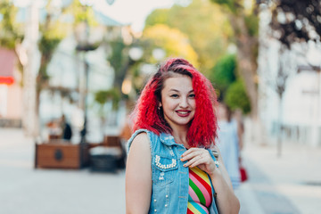 Stylish happy young woman in a bright striped sundress and a denim vest with red hair walks on the street. Portrait of a smiling girl